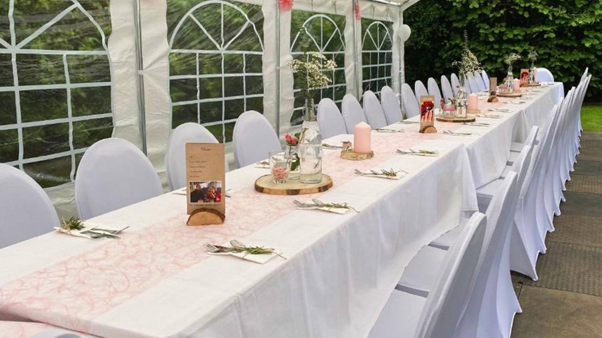Festlich gedeckte Tafel in einem Pavillonzelt mit weißen Stuhlhussen, Tischdeko und Blumen für eine Hochzeitsfeier